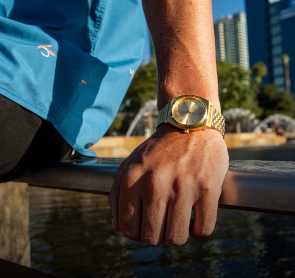 Person leaning against banister at waterfront wearing Nixon Time Teller in All Gold.